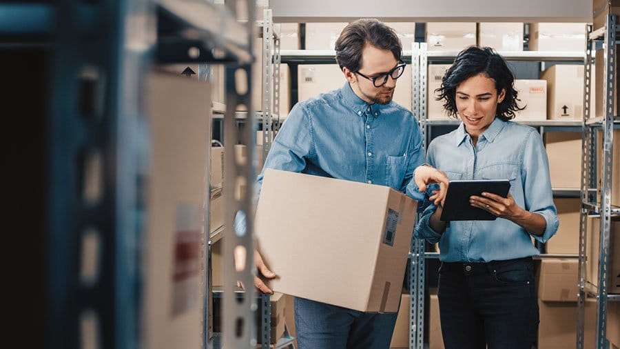 Two colleagues in a storage area, one holding a cardboard box and the other using a tablet, discussing inventory or logistics